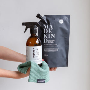 Photo of stretched out arms holding out an amber glass bathroom cleaner with a spray nozzle and a 1 litre refill pouch on a madekind emerald coloured cloth